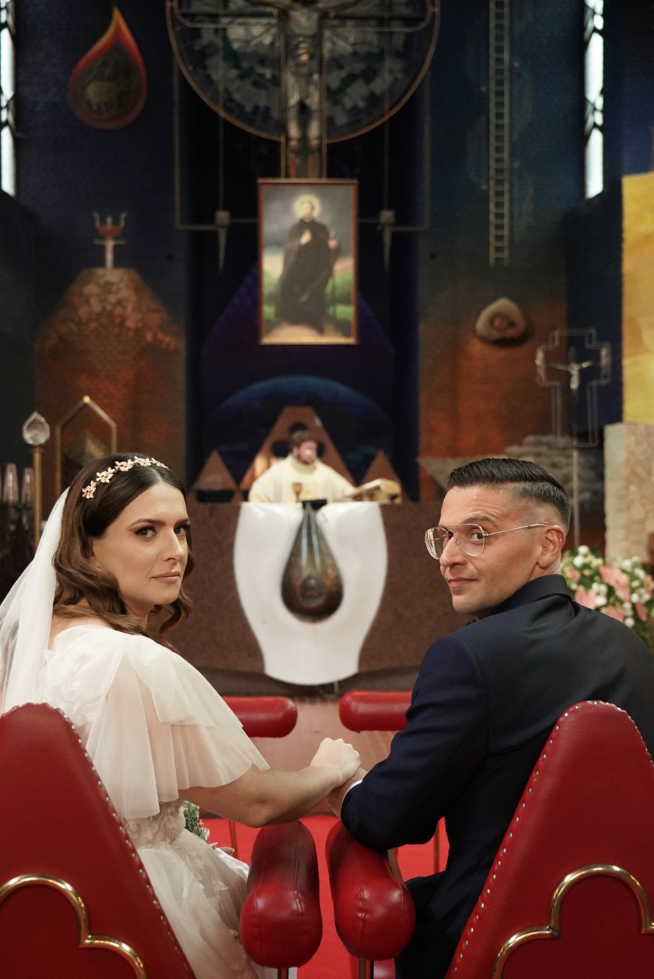 Para Młoda siedzi w kościele, trzymając się za ręce. W tle ołtarz i ksiądz. Uwagę przykuwa nowoczesny wystrój kościoła z motywami religijnymi.