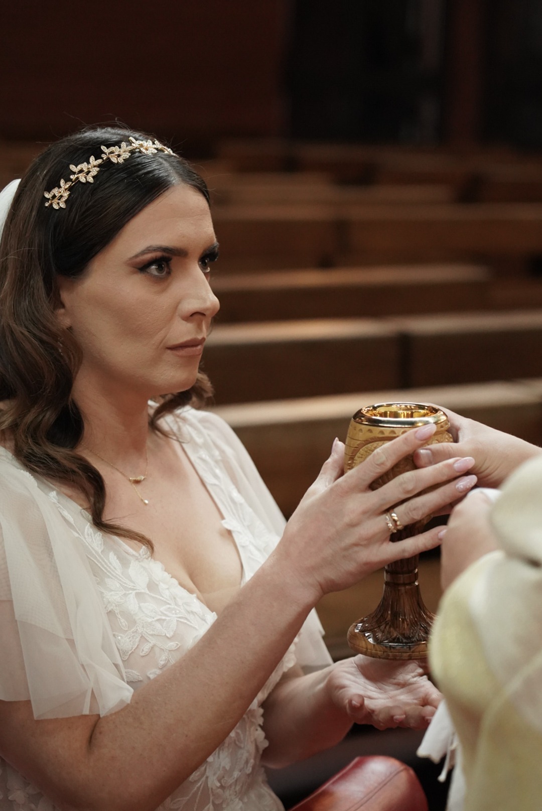 Panna młoda w białej sukni, z ozdobą we włosach, przyjmuje kielich podczas ceremonii ślubnej w kościele. W tle drewniane ławki.