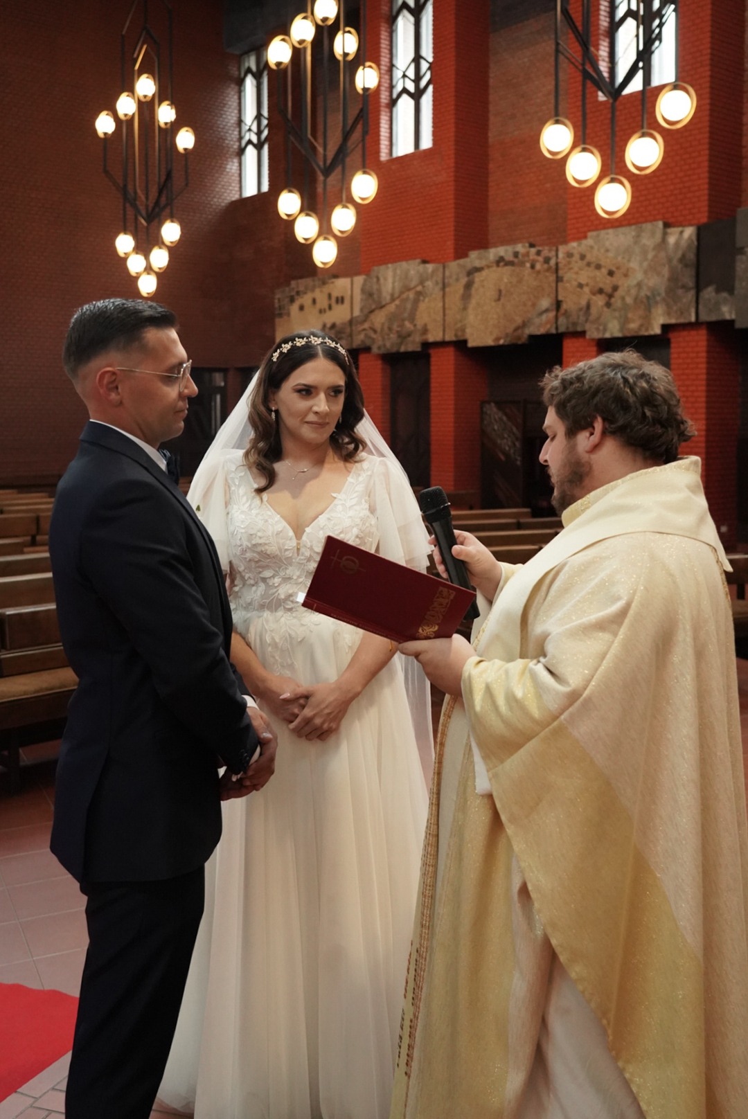 Uroczystość ślubna w kościele: para młoda stoi przed duchownym w bogato zdobionej szacie, który czyta z czerwonej księgi przy mikrofonie.