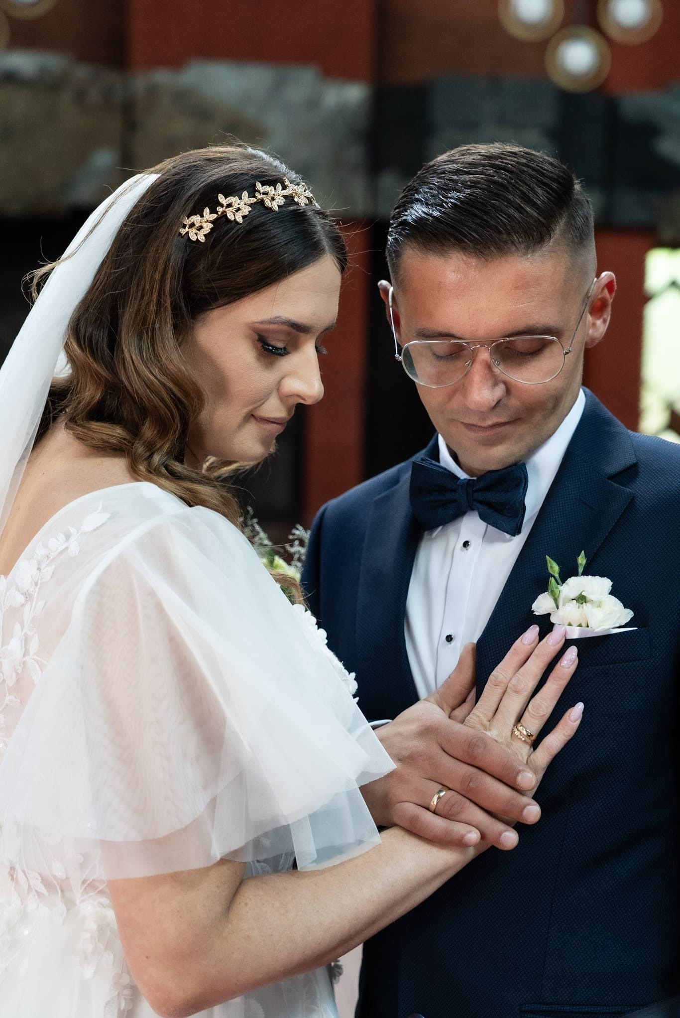 Para młoda wymienia obrączki podczas ceremonii ślubnej. Panna młoda w białej sukni i welonie, pan młody w granatowym garniturze i muszce. Delikatne światło podkreśla detale.