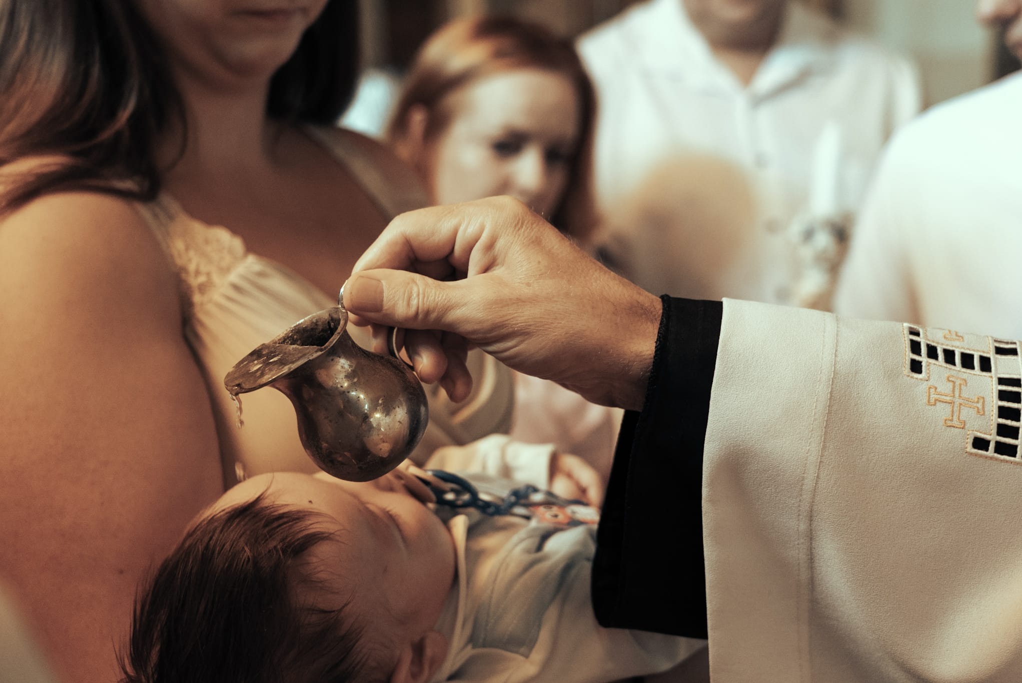 Chrzest dziecka: ksiądz polewa wodą głowę niemowlęcia z metalowego naczynia. W tle matka z dzieckiem i inni uczestnicy ceremonii. Uroczystość w kościele.