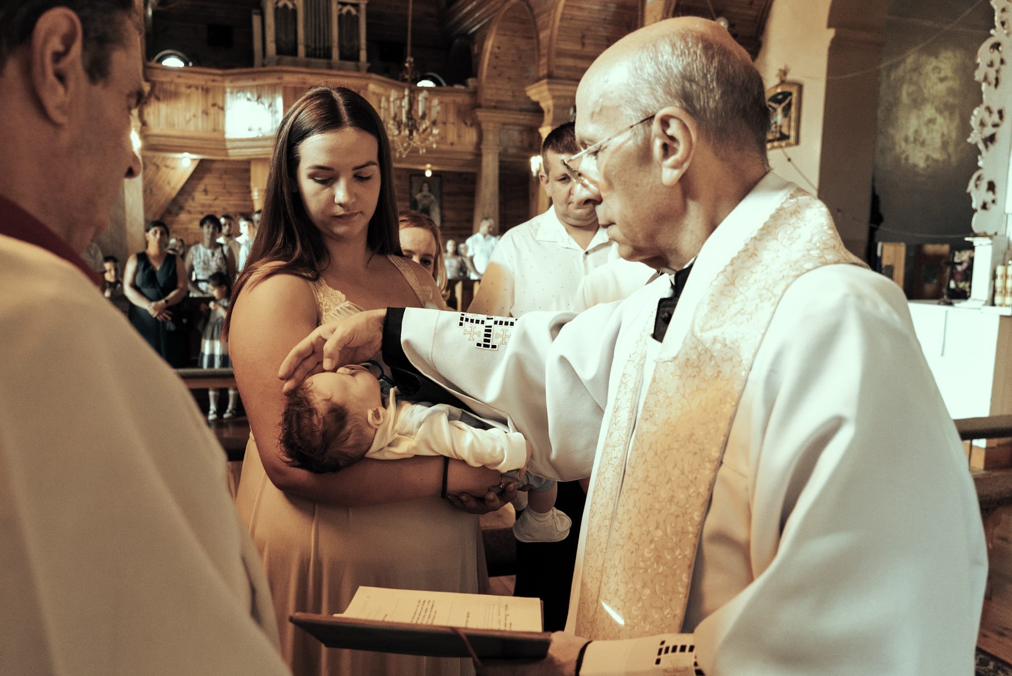 Chrzest dziecka w kościele z drewnianym wystrojem. Ksiądz w ornacie polewa główkę niemowlaka trzymanego przez matkę. W tle widoczni goście.