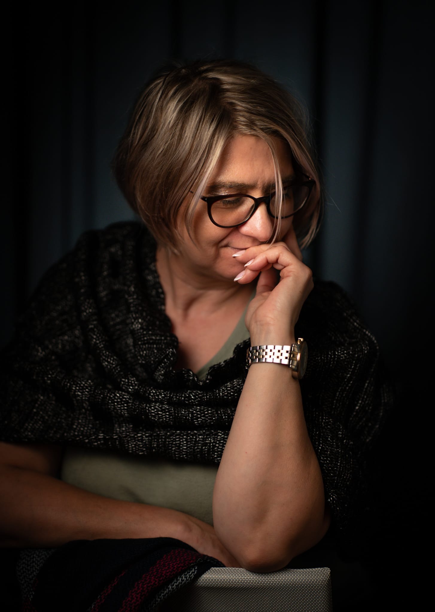 Portret kobiety w okularach, zamyślona, w ciemnym szalu, z ręką przy twarzy. Stonowana kolorystyka, miękkie światło, ciemne tło. Ujęcie od przodu, skupione na twarzy.