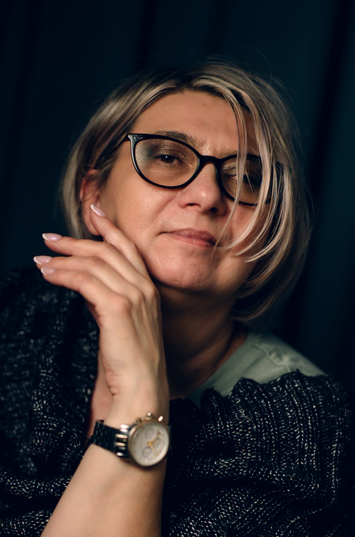 Portret kobiety w okularach, z blond włosami i zegarkiem na ręce, w ciemnym otoczeniu. Delikatny uśmiech i dłoń przy twarzy dodają subtelności.