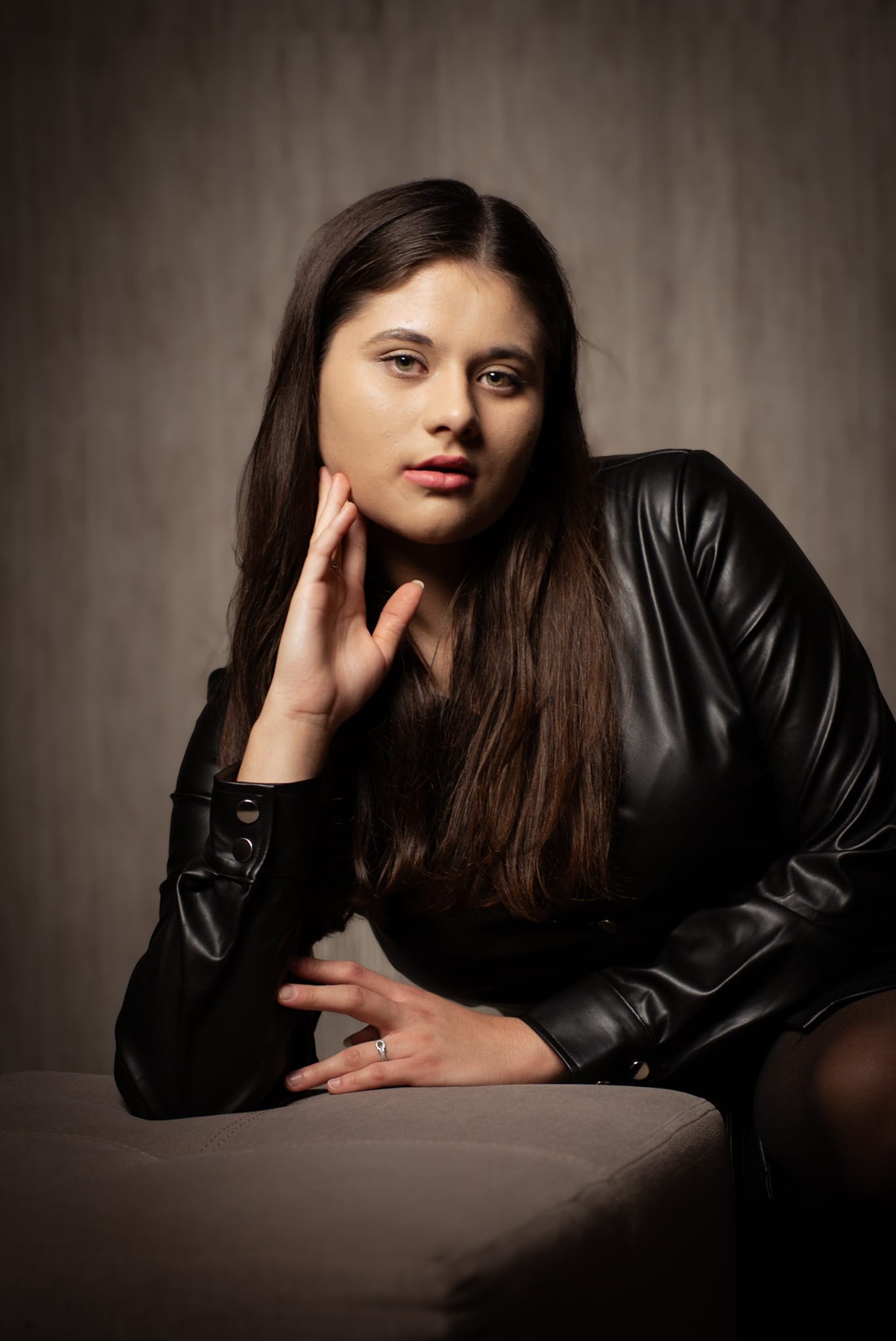 Portret kobiety w czarnej, skórzanej sukience, opierającej dłoń o policzek, z pierścionkiem na palcu, na neutralnym tle. Ujęcie z bliska, miękkie światło.