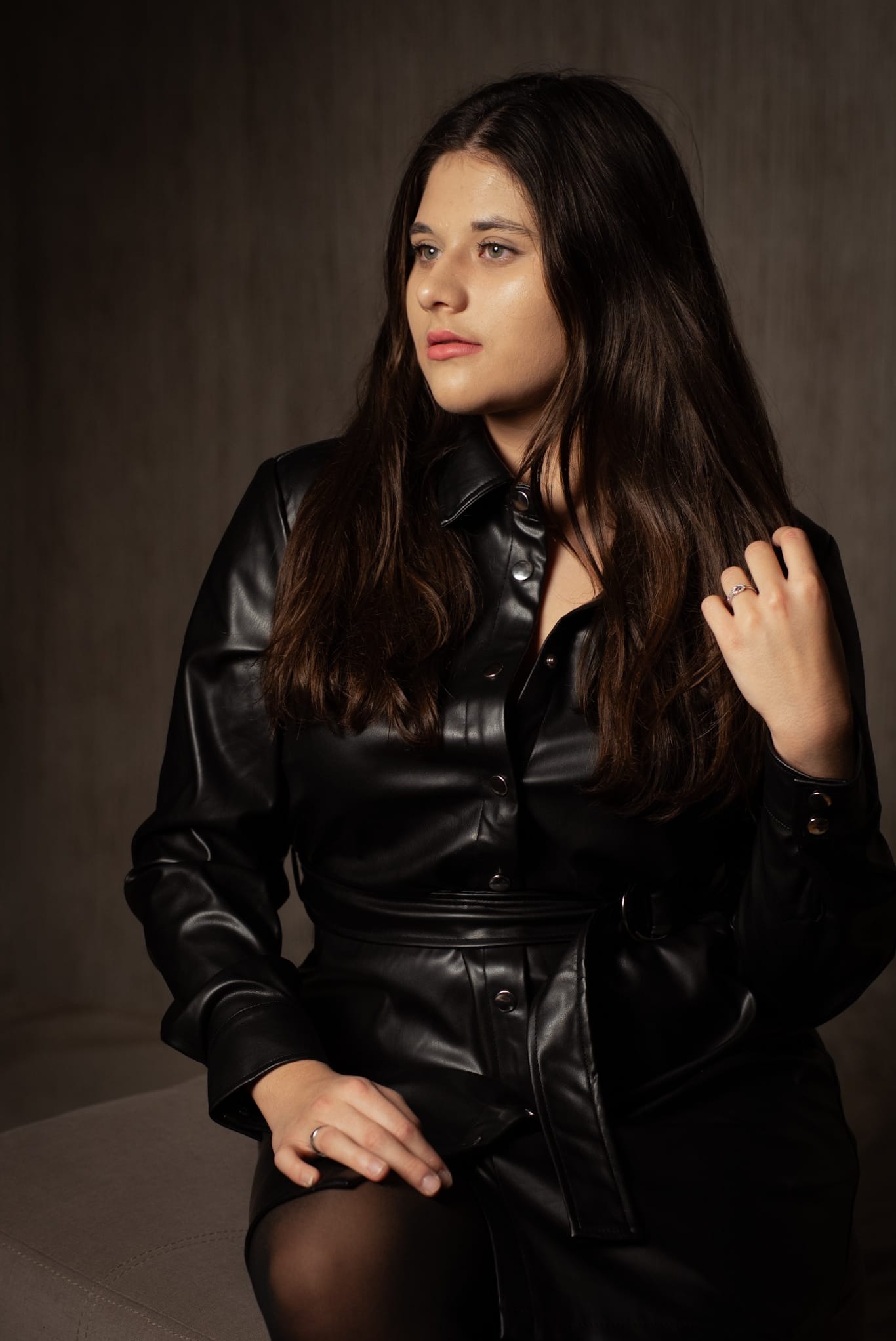 Portret kobiety w czarnej, skórzanej sukience na tle ciemnego, teksturowanego tła. Delikatne światło podkreśla rysy twarzy i fakturę materiału. Uwagę przyciąga pierścionek na palcu.