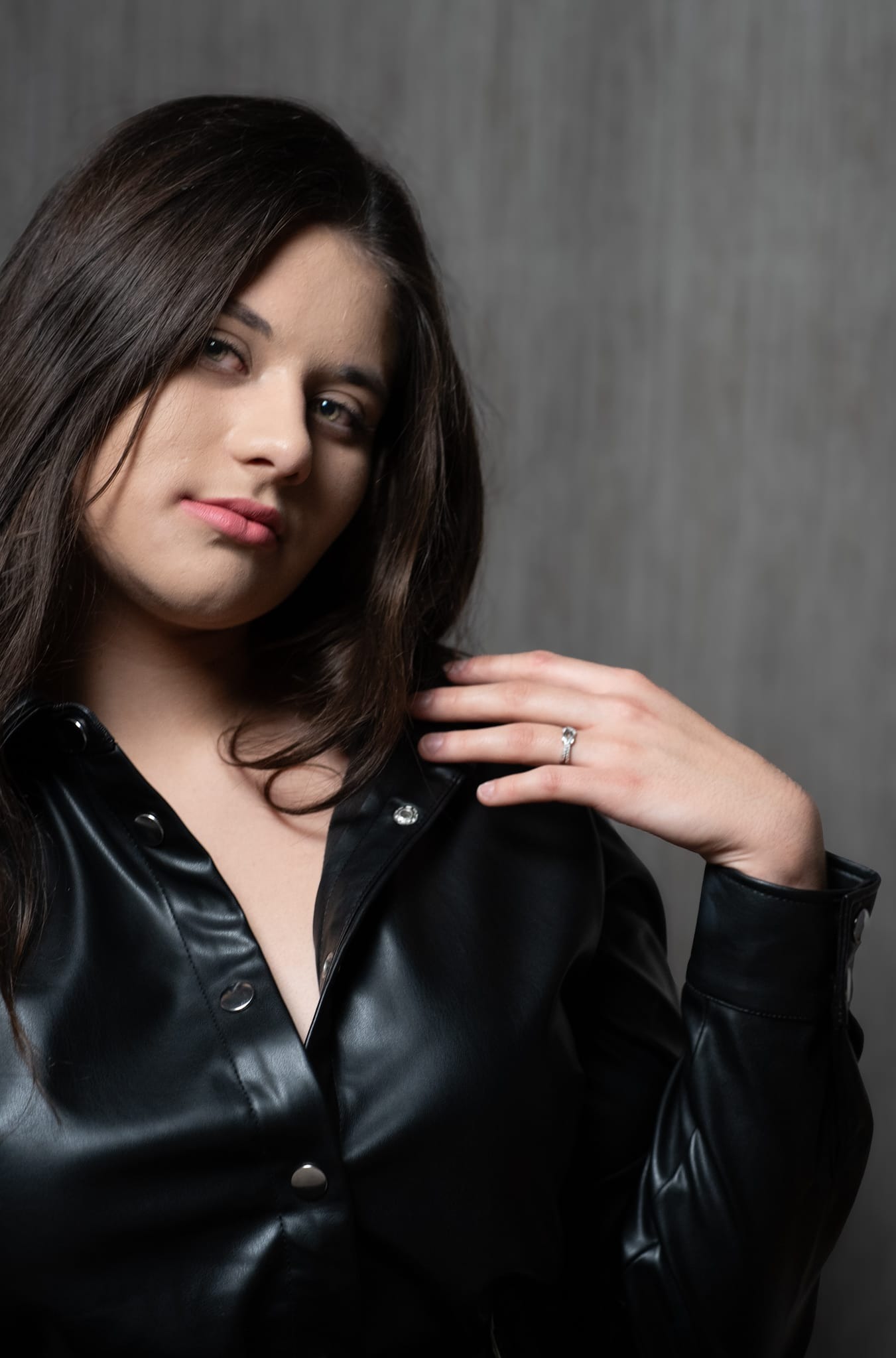 Portret kobiety w czarnej, skórzanej koszuli na tle szarej ściany. Na palcu widoczny pierścionek. Ujęcie z bliska, skupione na twarzy i dekolcie.