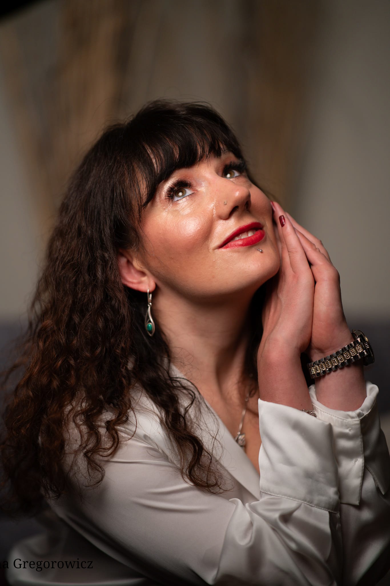 Portret kobiety z ciemnymi kręconymi włosami, w białej koszuli, z biżuterią, patrzy w górę, dłonie złożone blisko twarzy. Usta pomalowane czerwoną szminką.