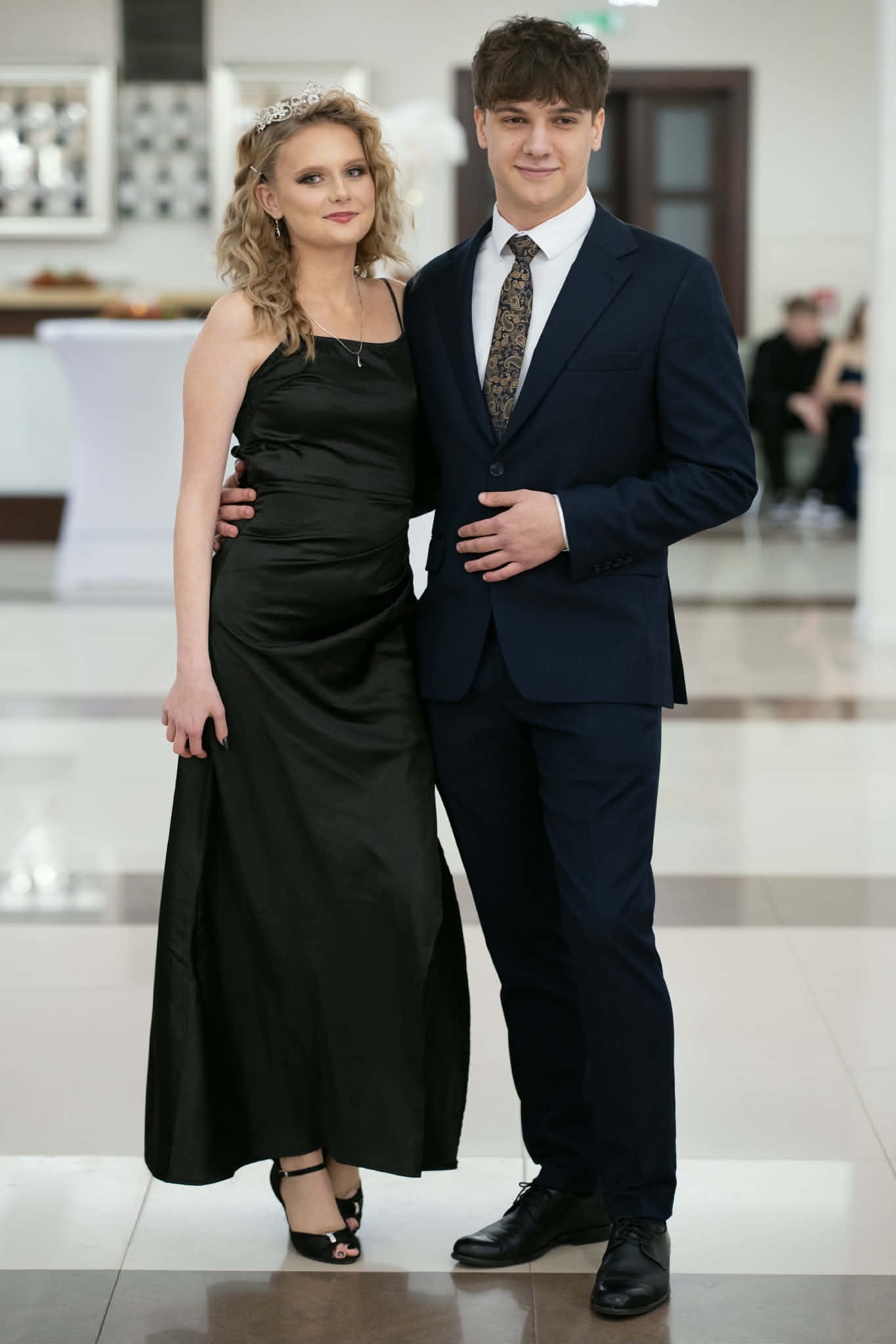 Elegancka para: blondwłosa kobieta w czarnej sukni i mężczyzna w granatowym garniturze, pozujący na jasnym tle. Ujęcie portretowe, pełne wdzięku i szyku.