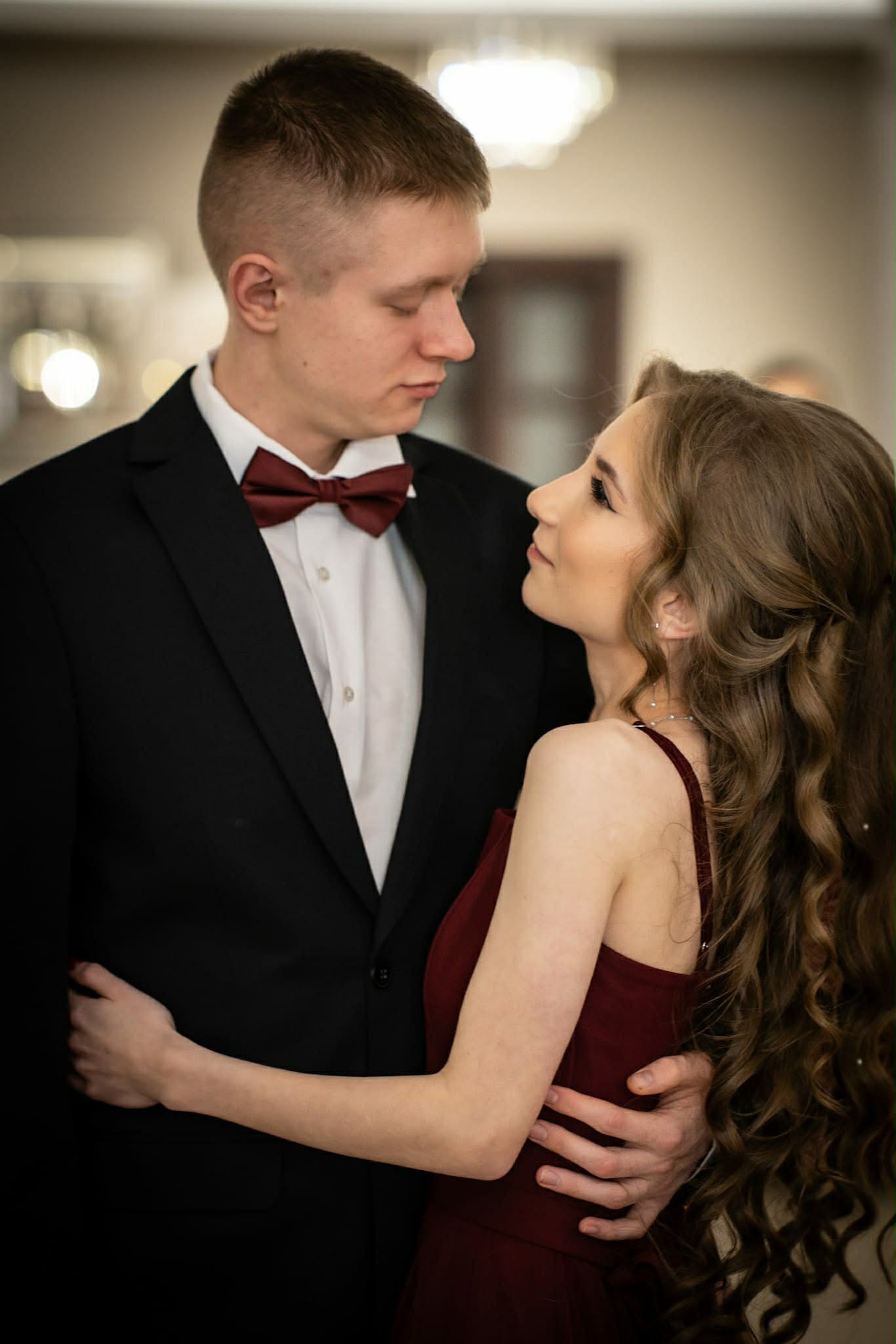 Elegancka para: mężczyzna w smokingu z bordową muchą i kobieta w bordowej sukience, obejmujący się i patrzący na siebie z czułością w neutralnym wnętrzu.