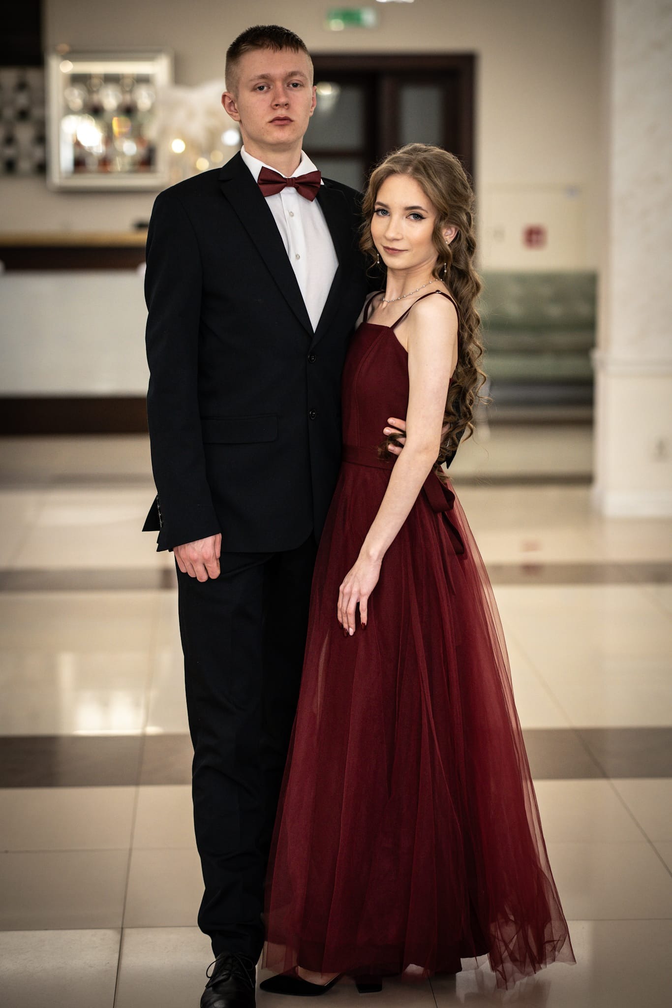 Elegancka para: mężczyzna w czarnym garniturze i bordowej muszce oraz kobieta w długiej, bordowej sukni, pozują w jasnym wnętrzu z marmurową podłogą.