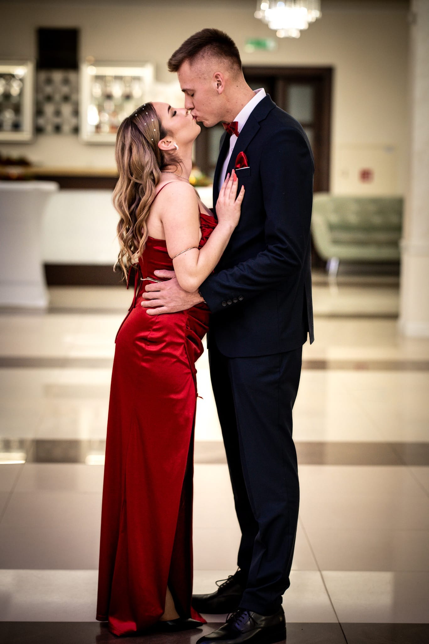 Elegancka para w pocałunku: kobieta w czerwonej sukni, mężczyzna w granatowym garniturze z czerwoną muchą, w jasnym wnętrzu z elementami dekoracyjnymi.