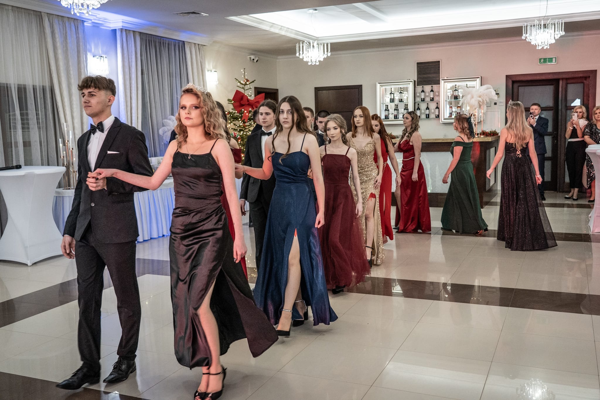 Uczestnicy studniówki w eleganckich strojach, defilujący w sali balowej z dekoracjami świątecznymi w tle. Mężczyzna w garniturze, kobiety w długich sukniach.