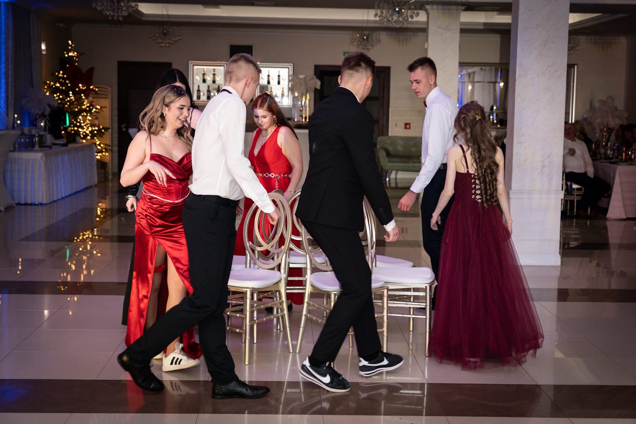 Grupa młodych ludzi w eleganckich strojach, w tym czerwone suknie, w przestronnym, oświetlonym wnętrzu z dekoracjami świątecznymi w tle. Parkiet z połyskiem.