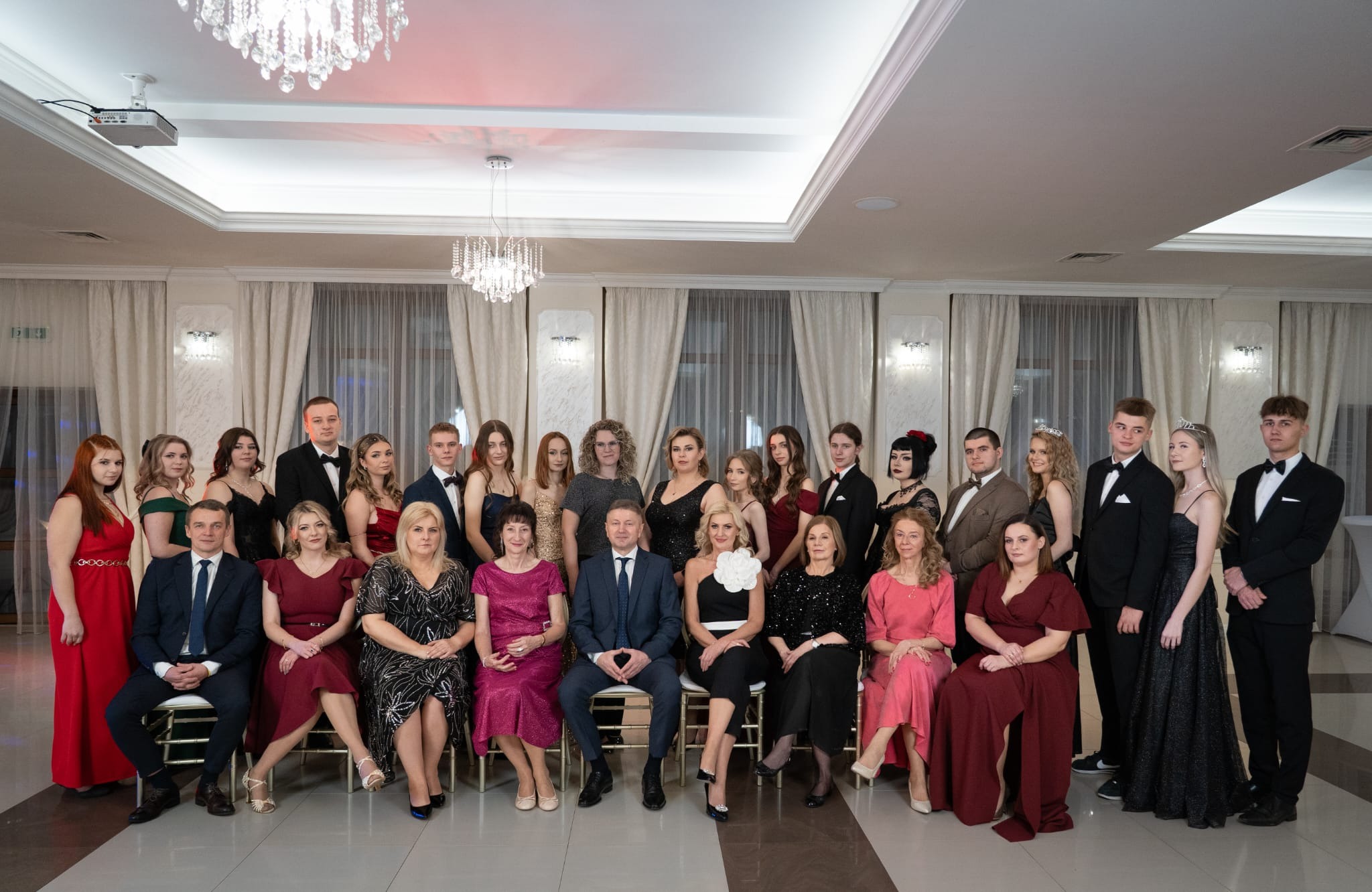Grupowe, pozowane zdjęcie uczestników balu w eleganckich strojach wieczorowych, w przestronnym, oświetlonym sali balowej z dekoracjami i żyrandolami.