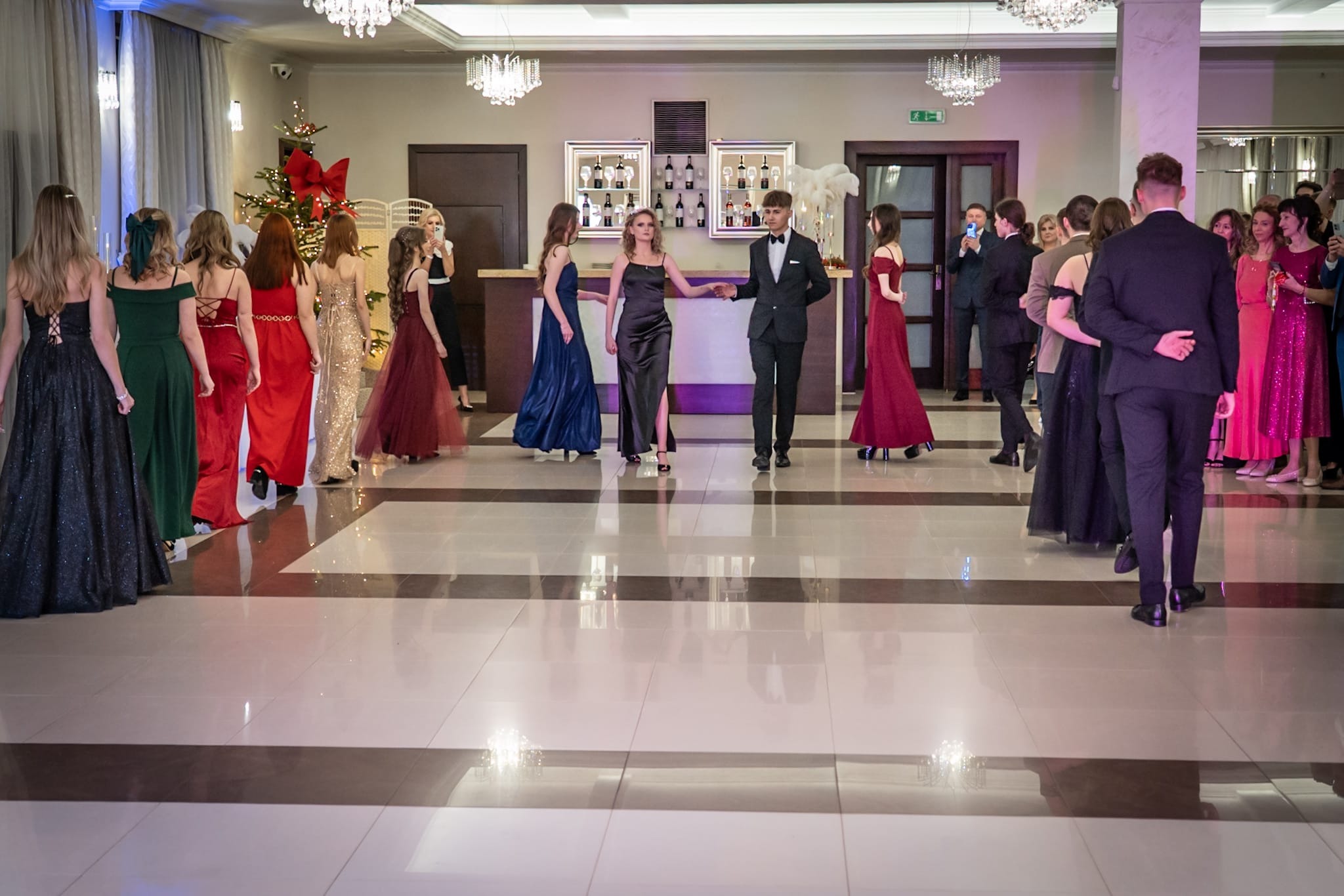 Uroczysta sala balowa z młodymi ludźmi w eleganckich strojach, przygotowującymi się do poloneza. W tle bar i dekoracje świąteczne.