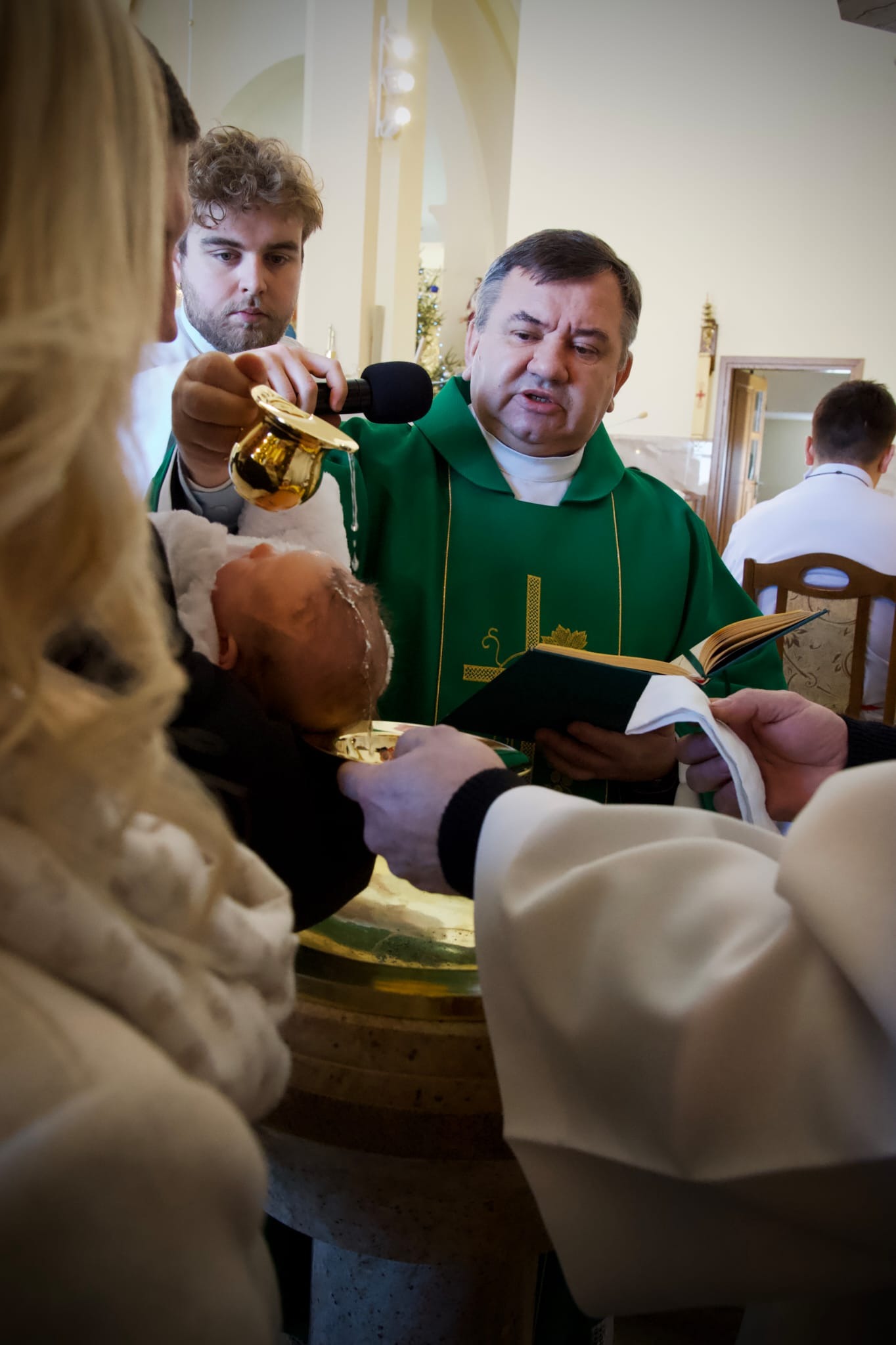 Kadr z ceremonii chrztu: ksiądz w zielonej szacie polewa wodą głowę dziecka, asystują rodzice. W tle wnętrze kościoła z delikatnym oświetleniem.