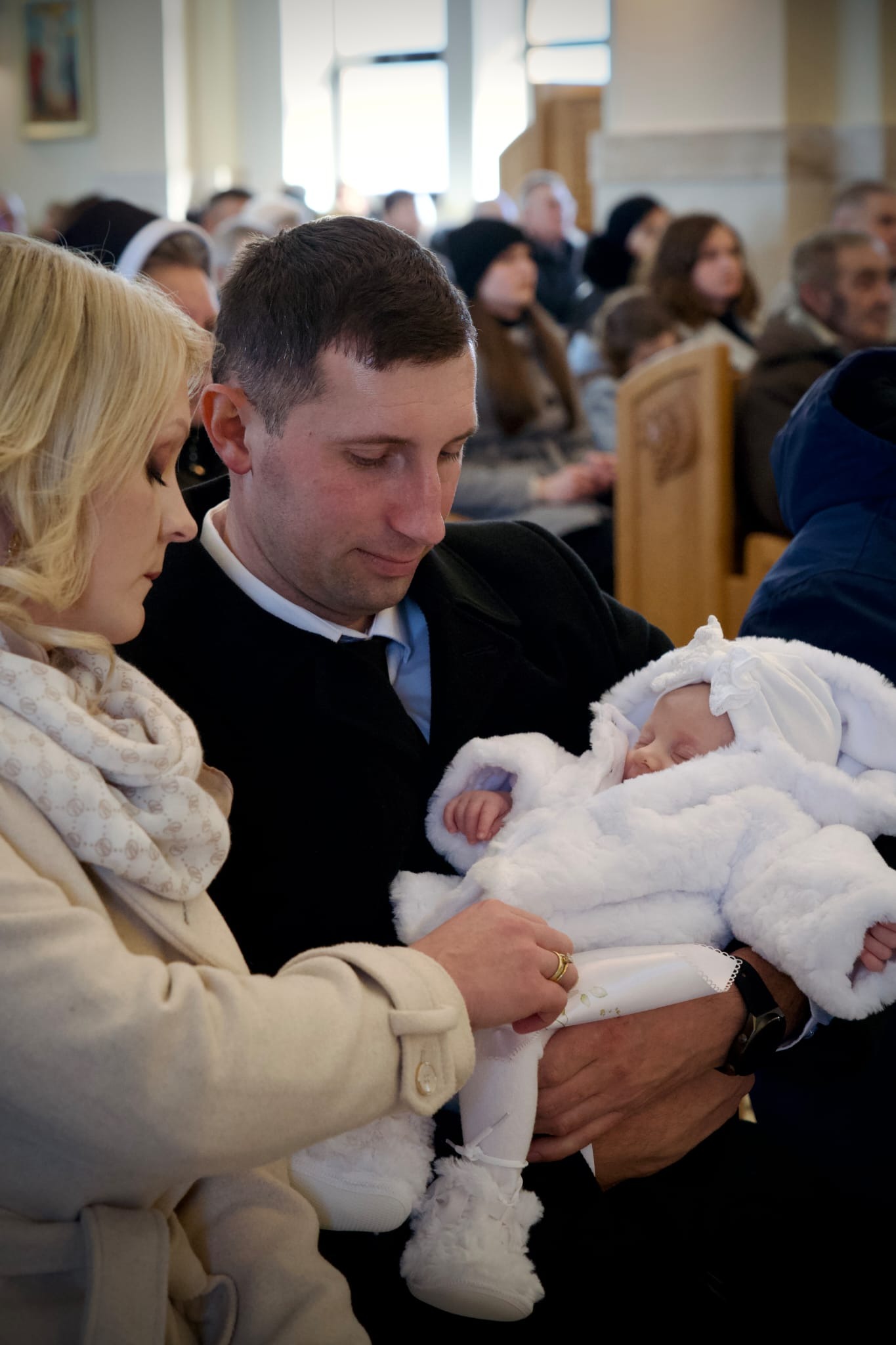 Rodzice z śpiącym niemowlęciem w białym ubranku podczas uroczystości w kościele. Mama delikatnie poprawia ubranko dziecka, w tle widoczni goście.