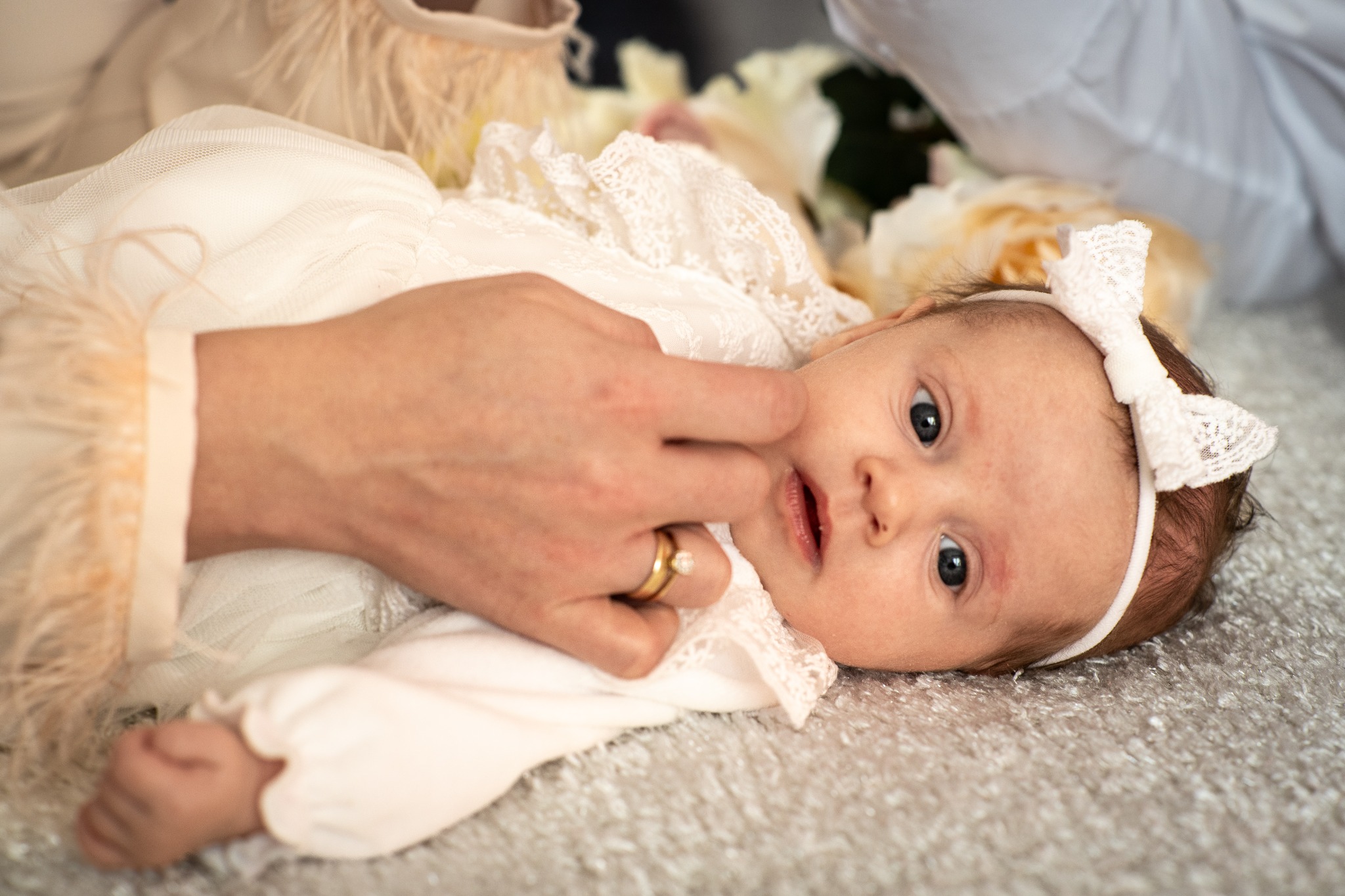 Urocze niemowlę w białej sukience i opasce z kokardą, leżące na miękkim dywanie, delikatnie dotykane przez dłoń z pierścionkiem. Kwiaty w tle.