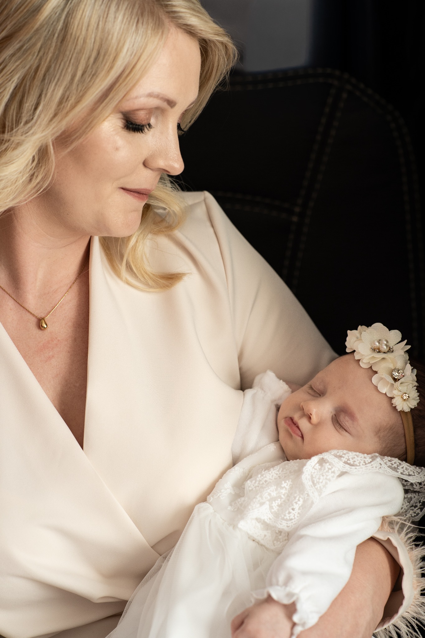 Portret matki z śpiącym noworodkiem w białej sukience i opasce z kwiatami. Delikatne oświetlenie, profil matki, ciemne tło. Ujęcie z bliska, skupienie na twarzach.