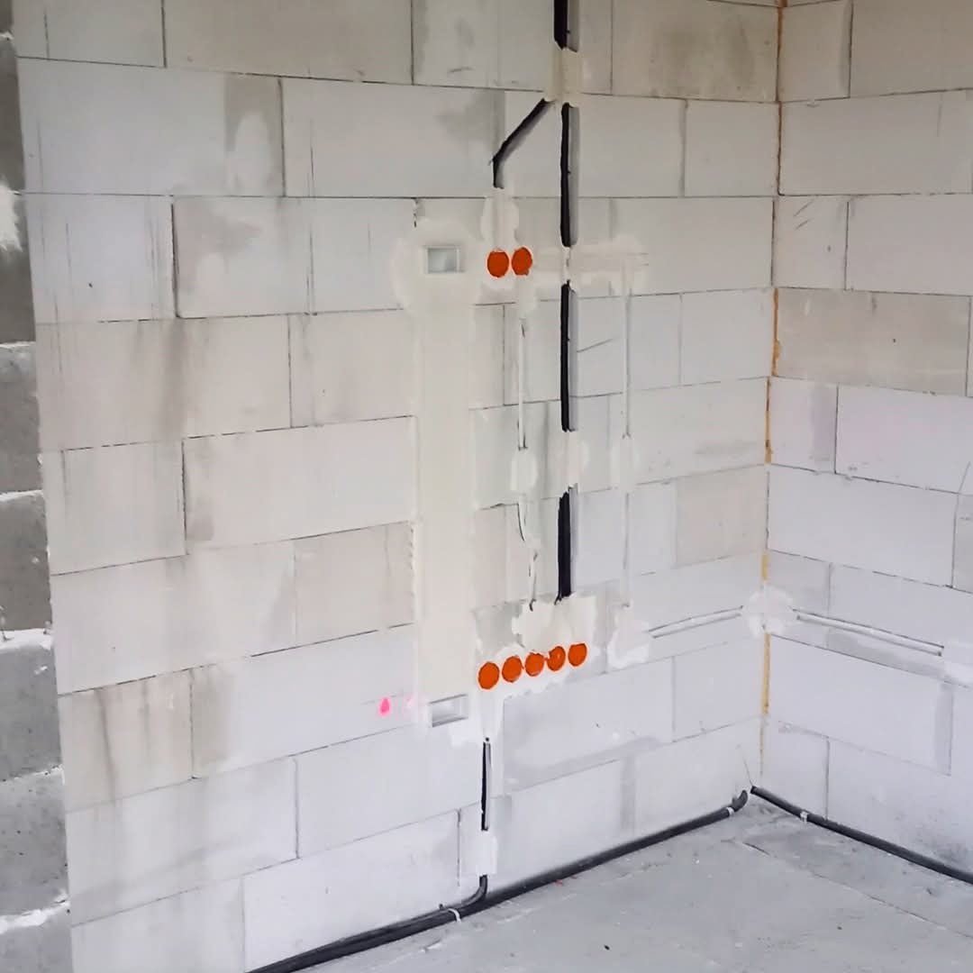 Surowe ściany z betonu komórkowego z pionowo rozprowadzonymi przewodami elektrycznymi, pomalowanymi na czarno i zabezpieczonymi gipsem. Widoczne pomarańczowe puszki.