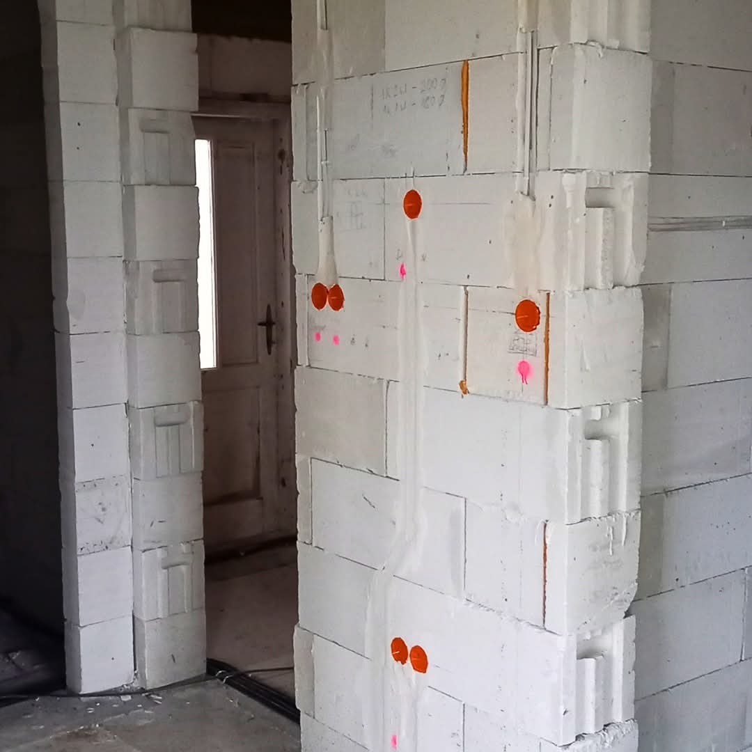 Białe ściany z betonu komórkowego w trakcie instalacji elektrycznej. Widoczne kable w ścianach, oznaczone punktami w kolorze pomarańczowym i różowym. Drzwi w tle.