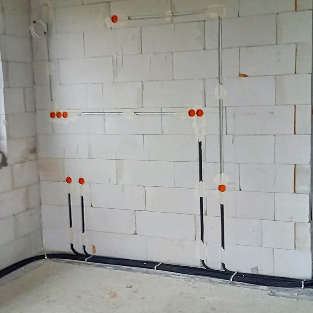 Surowa ściana z białego bloczka z widoczną, częściowo zamaskowaną instalacją elektryczną w peszlu i puszkami. Widok na etapie prac wykończeniowych.