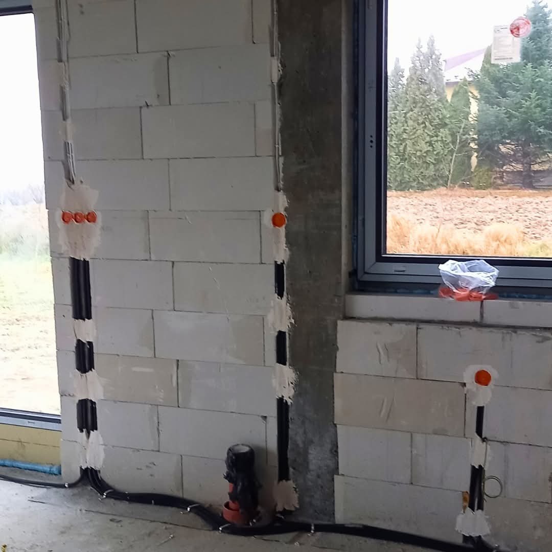 Wewnętrzna instalacja elektryczna w surowym budynku z białych bloczków, widoczne kable w czarnych peszlach i pomarańczowe puszki pod gniazdka, okno z widokiem na zewnątrz.