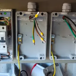 Otwarta skrzynka elektryczna z bezpiecznikami i podłączonymi przewodami w kolorach: zielonym, żółtym, brązowym i szarym. Widoczne oznaczenia i naklejki na obudowie.