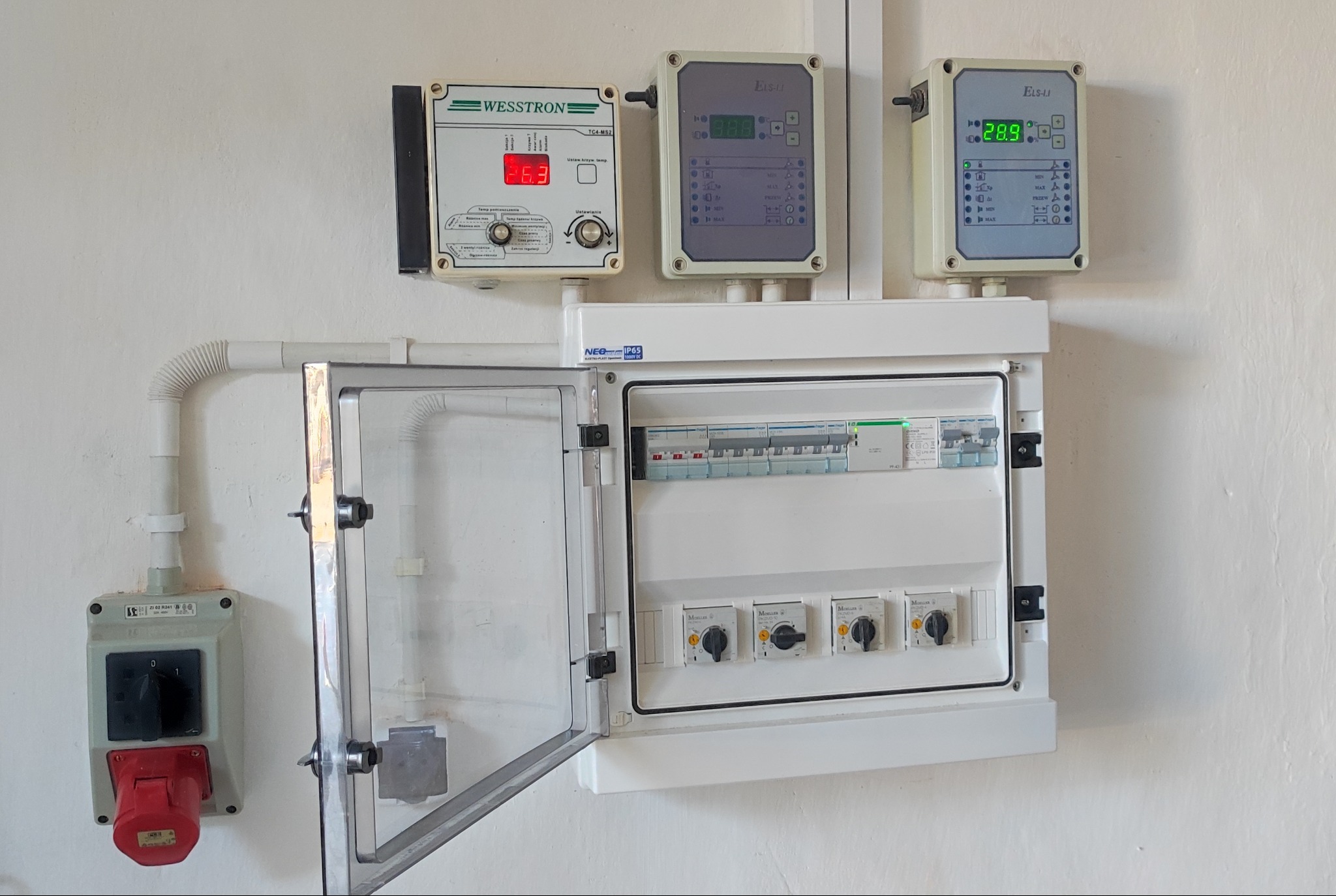 Rozdzielnica elektryczna z otwartymi drzwiczkami, zabezpieczenia, wyłączniki, sterowniki i gniazdo siłowe na białej ścianie. Widoczne elementy instalacji elektrycznej.