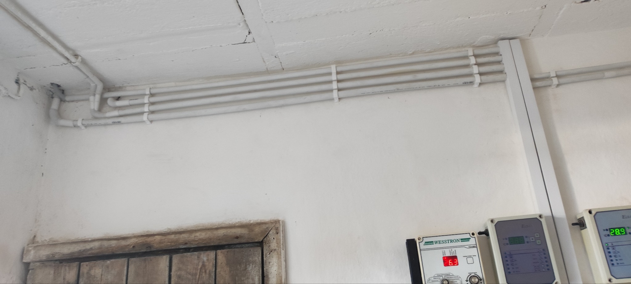 Białe rury instalacyjne przymocowane do sufitu i ściany, z widocznymi elementami sterującymi na ścianie. Widoczne okablowanie i złącza.