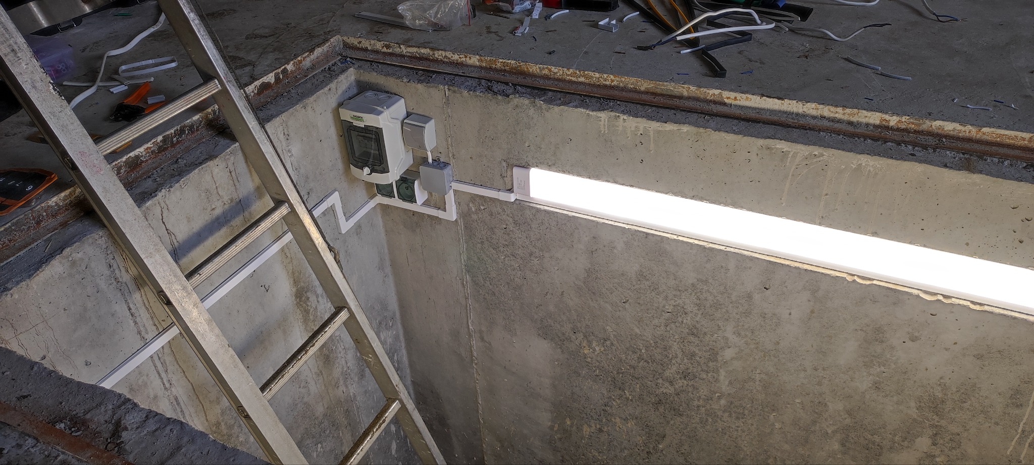 Betonowy szyb z zamontowaną lampą LED i skrzynką elektryczną. Widoczna drabina i narzędzia. Oświetlenie w surowym, przemysłowym stylu.