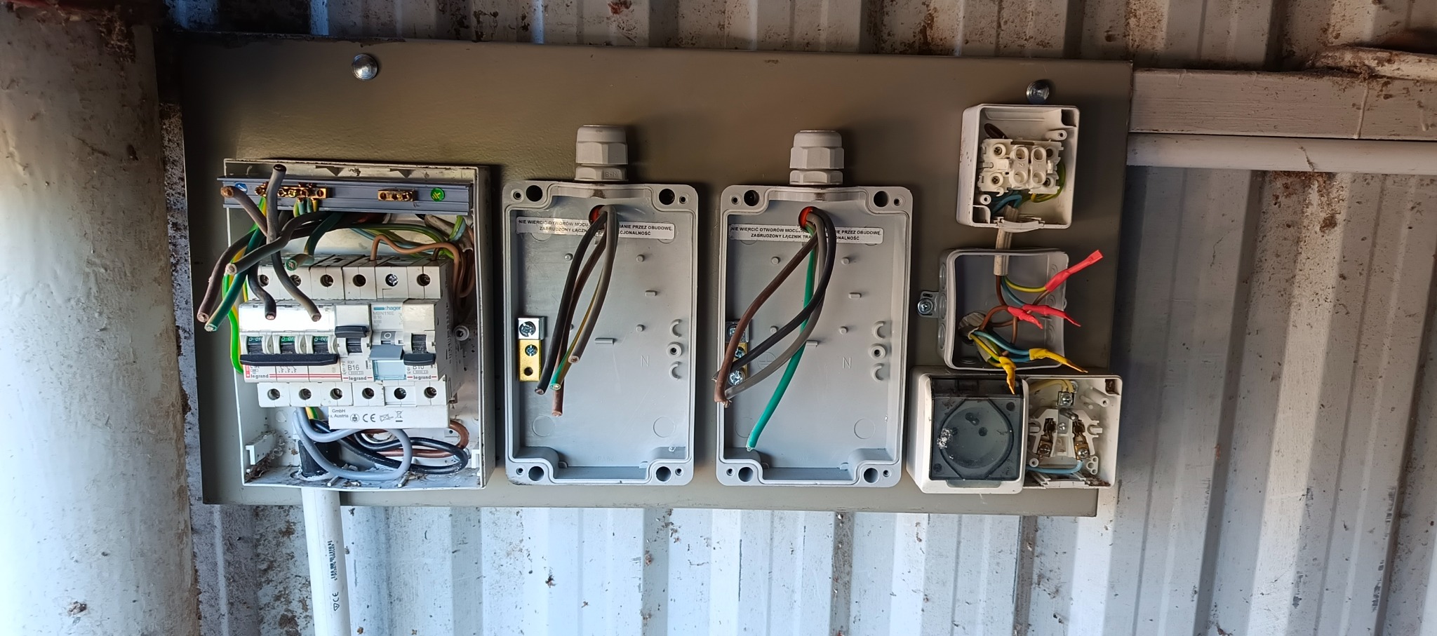 Rozdzielnica elektryczna z otwartymi puszkami i podłączonymi przewodami, zamontowana na ścianie z blachy falistej. Widoczne bezpieczniki i gniazda.