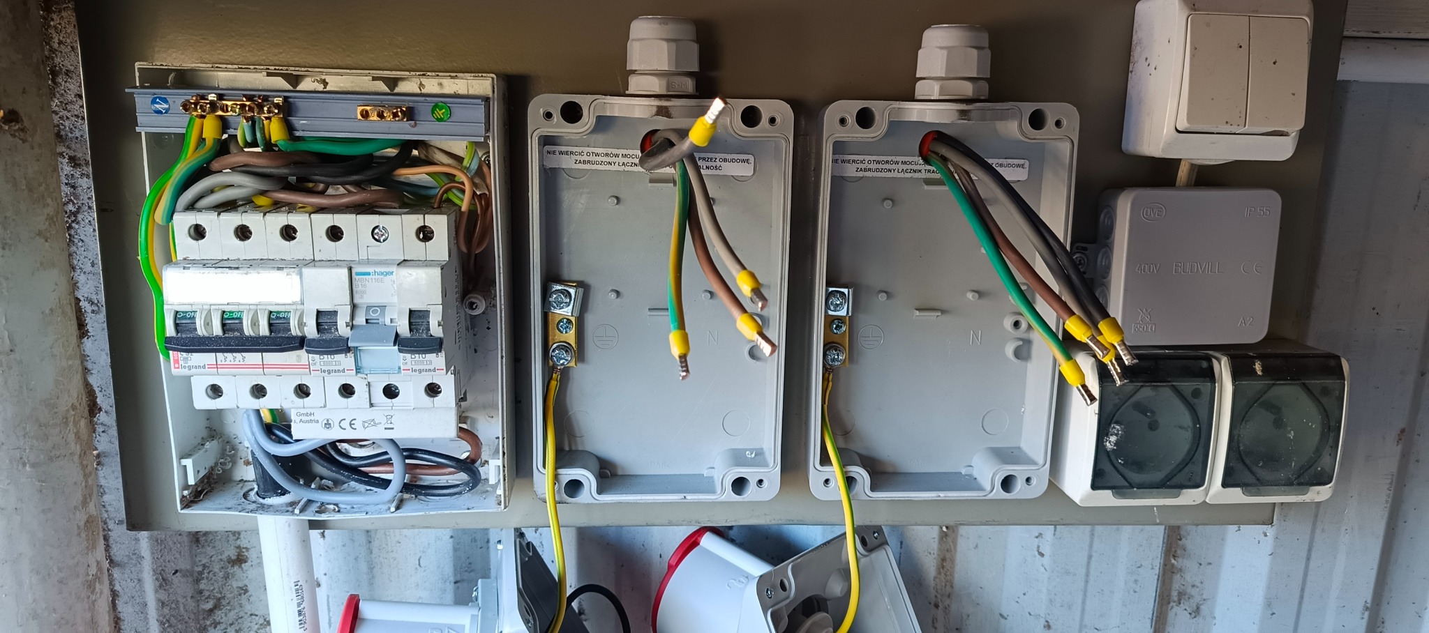 Otwarta skrzynka elektryczna z bezpiecznikami i podłączonymi przewodami w kolorach: zielonym, żółtym, brązowym i szarym. Widoczne oznaczenia i naklejki na obudowie.