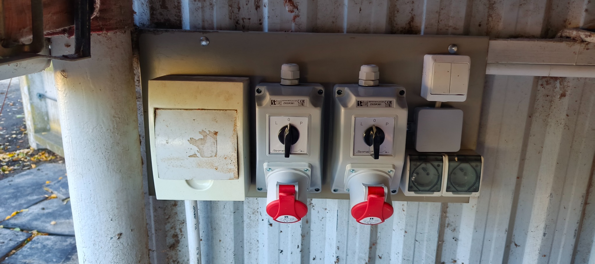 Szare rozłączniki elektryczne z czerwonymi wtykami zamontowane na metalowej płycie z widocznymi śladami użytkowania i zabrudzeniami, w tle blaszana ściana.