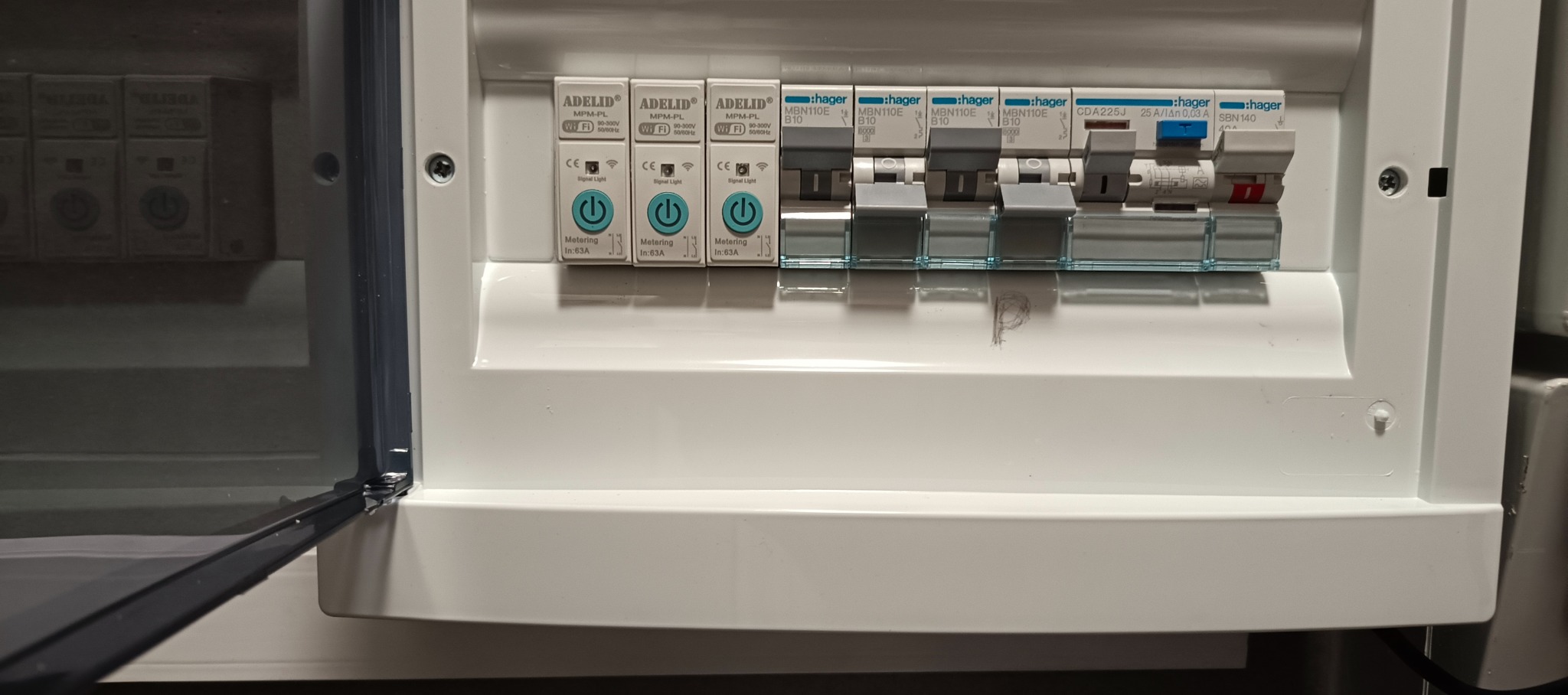 Otwarta, biała skrzynka elektryczna z zainstalowanymi bezpiecznikami Hager i modułami ADELID MPM-PL z włącznikami. Widoczne oznaczenia i parametry urządzeń.