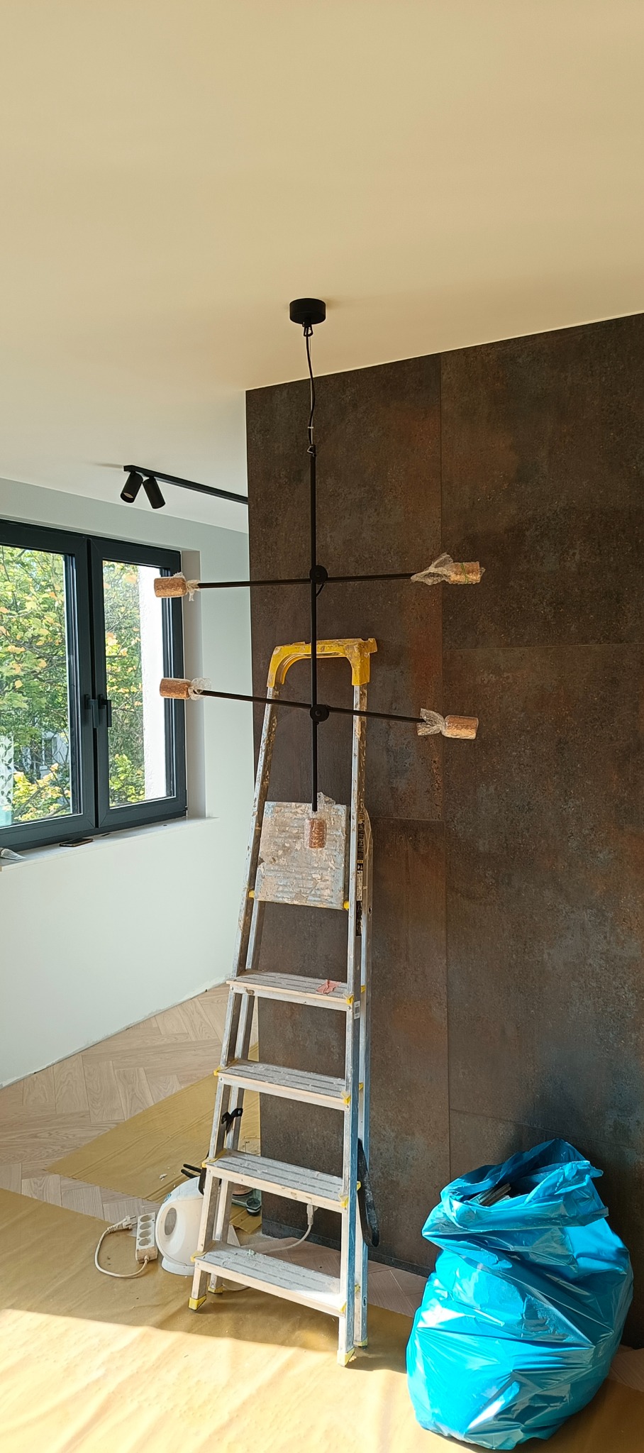 Nowoczesna czarna lampa wisząca z drewnianymi elementami, zamontowana na suficie. Pod nią drabina, a obok niebieski worek na śmieci. Widoczne również reflektory.