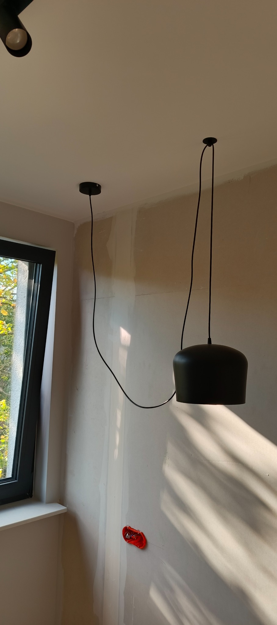 Czarna lampa wisząca z długim przewodem zamontowana na suficie w pokoju z oknem i przygotowaną instalacją elektryczną w ścianie.