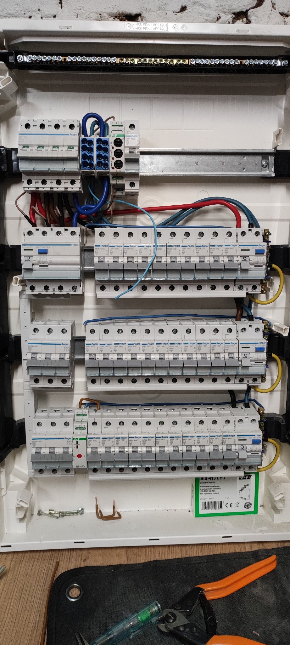 Rozdzielnica elektryczna Hager z zainstalowanymi bezpiecznikami i przewodami. Widoczne narzędzia elektryczne. Biała obudowa rozdzielnicy na tle ściany.