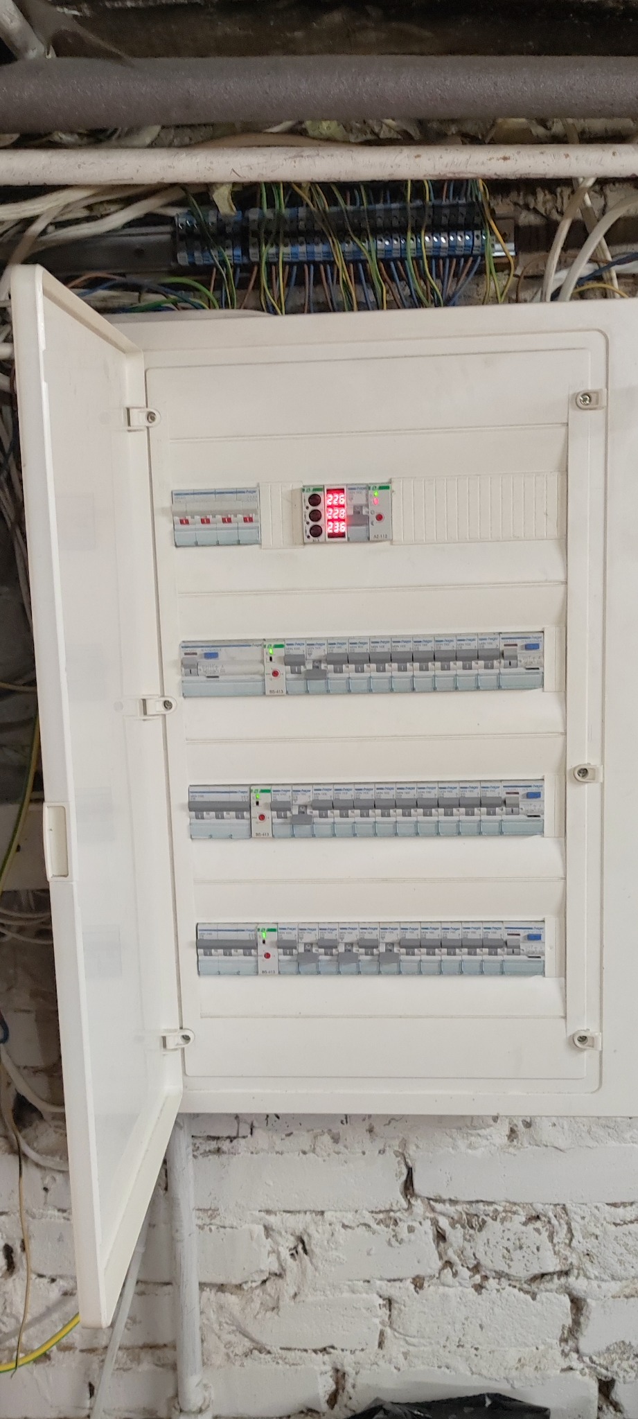 Otwarta skrzynka elektryczna z bezpiecznikami i wyświetlaczem napięcia na białej ścianie z cegły, widoczne kable u góry.