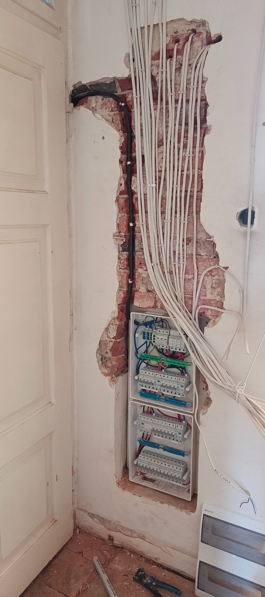 Rozdzielnica elektryczna w trakcie montażu w ścianie z odsłoniętą cegłą, widoczne kable i narzędzia. Prace instalacyjne w starym budownictwie.