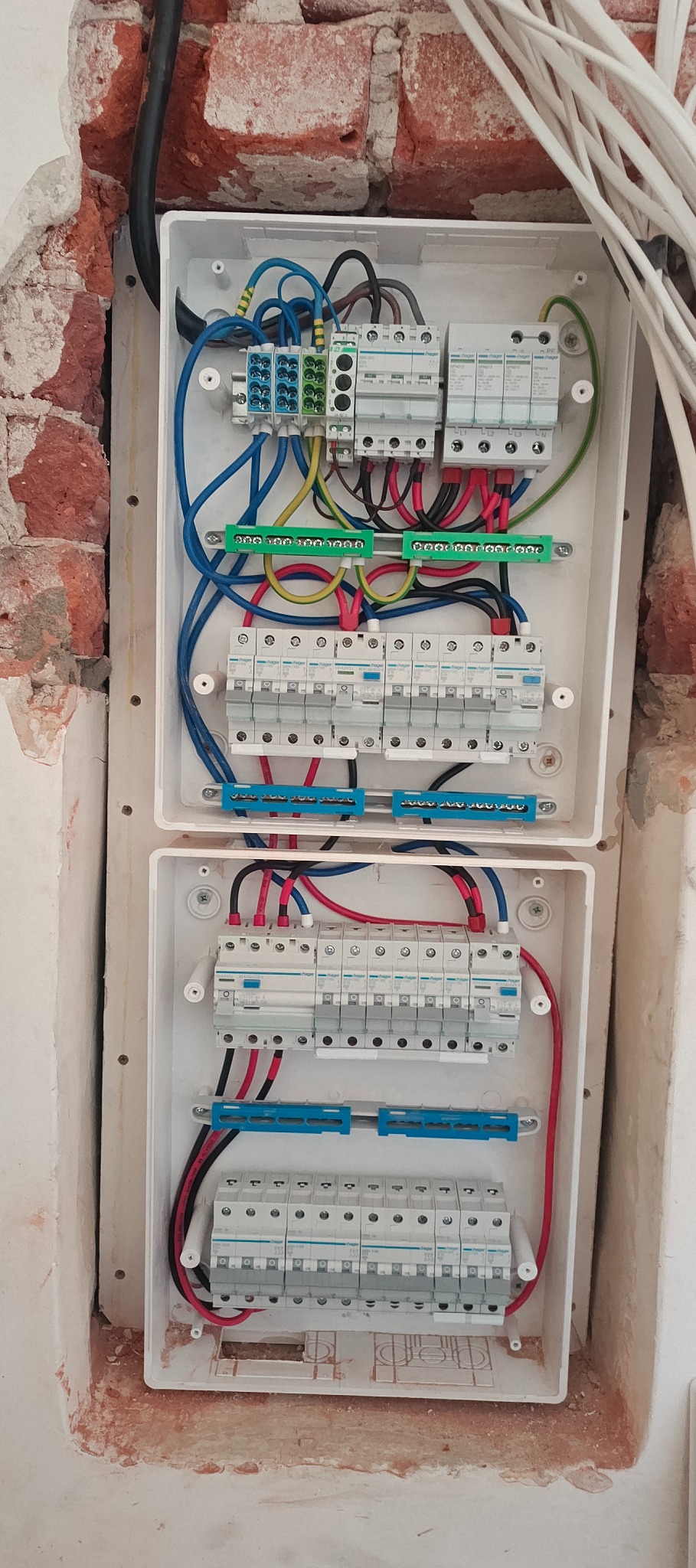 Dwie białe skrzynki z zainstalowanymi bezpiecznikami i okablowaniem, zamontowane w ścianie z odsłoniętą cegłą. Widoczne przewody w różnych kolorach.