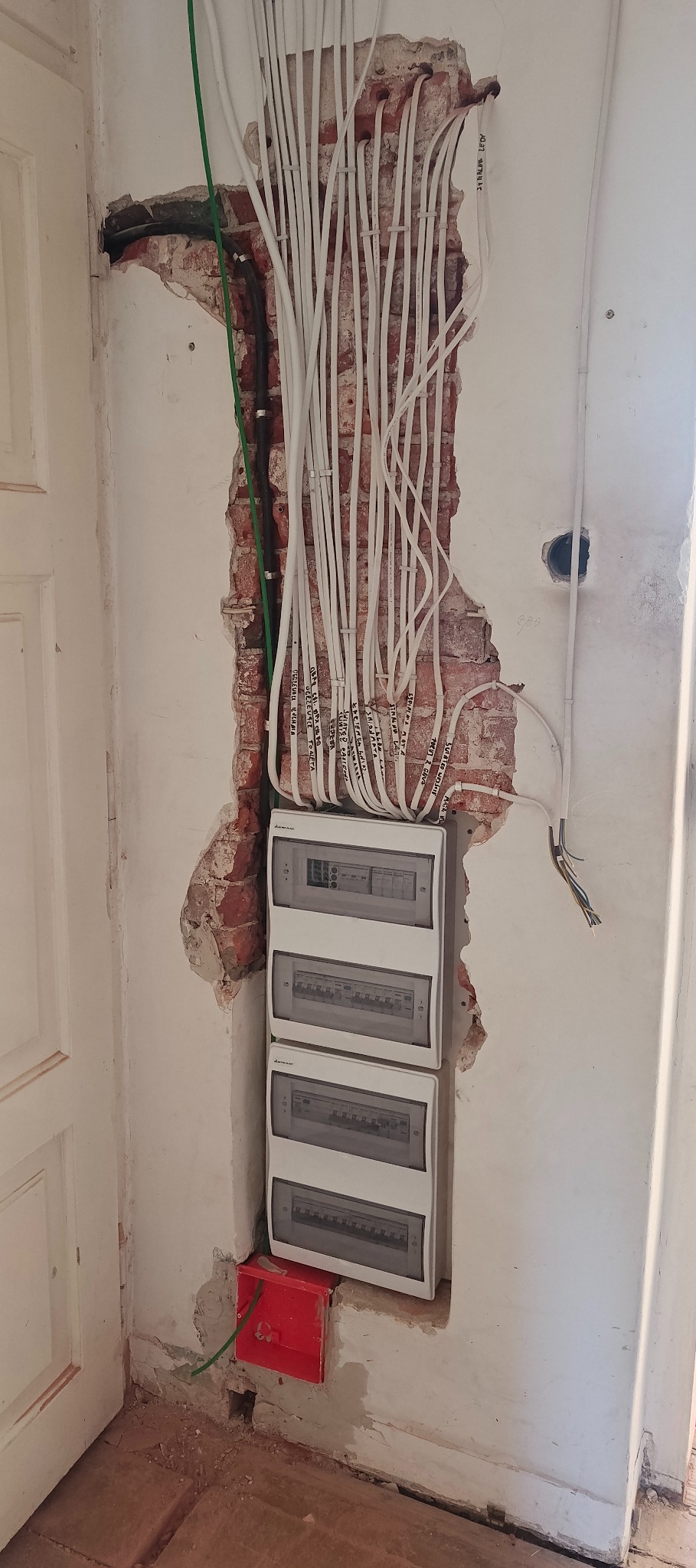 Rozdzielnia elektryczna z przewodami w ścianie z odsłoniętą cegłą. Przewody opisane, prowadzone wzdłuż ściany. Widoczna skrzynka instalacyjna i fragment drzwi.