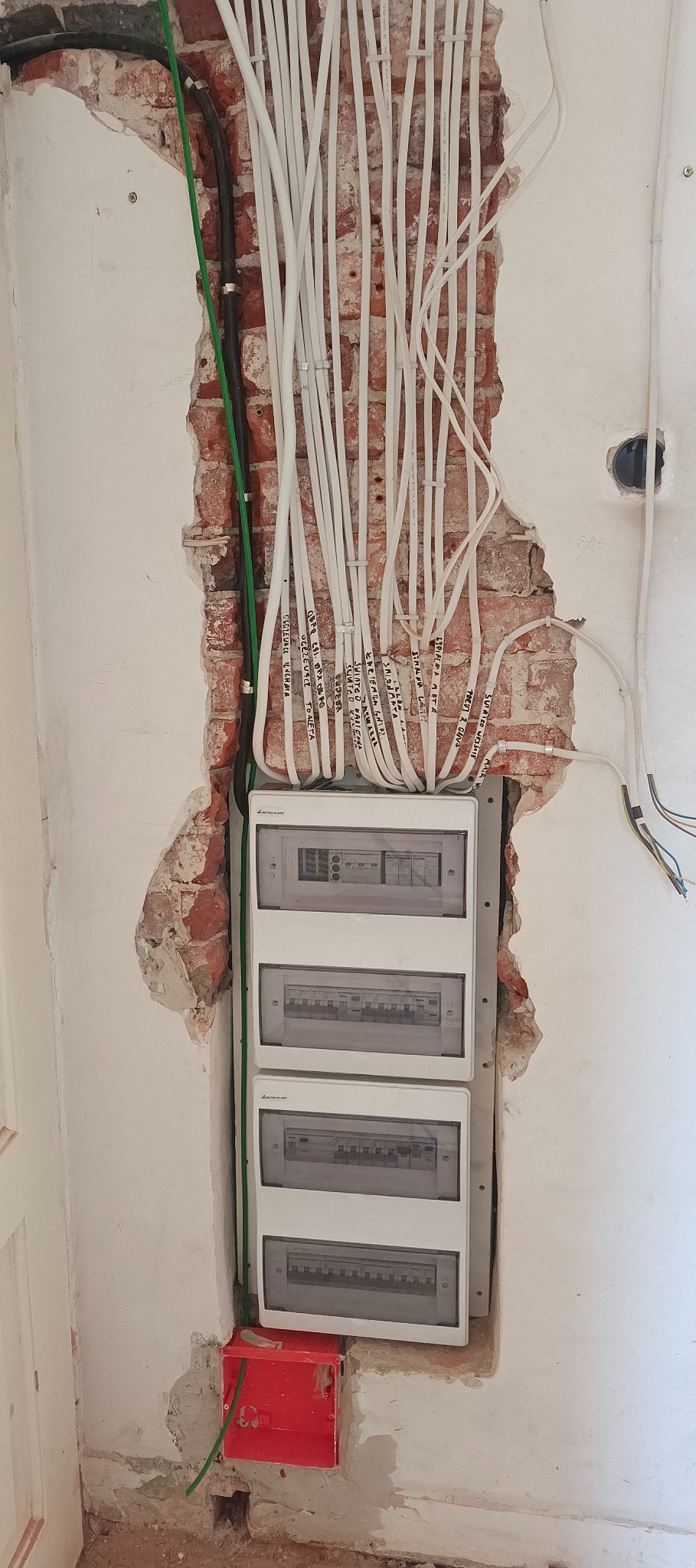 Rozdzielnia elektryczna z licznymi przewodami w ścianie z odsłoniętą cegłą. Widoczne kable opisane markerem. Czerwona puszka instalacyjna na dole. Etap prac instalacyjnych.