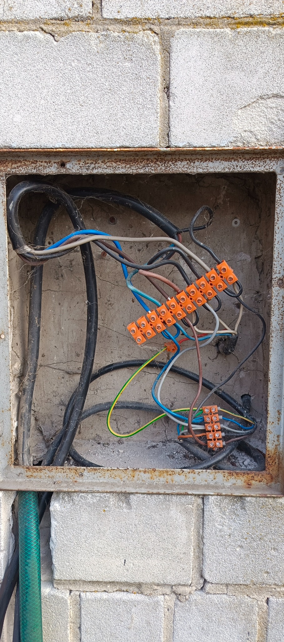 Otwarta, zardzewiała puszka elektryczna w murze z widocznymi przewodami i pomarańczowymi złączkami Wago. Widoczne pajęczyny i zabrudzenia.