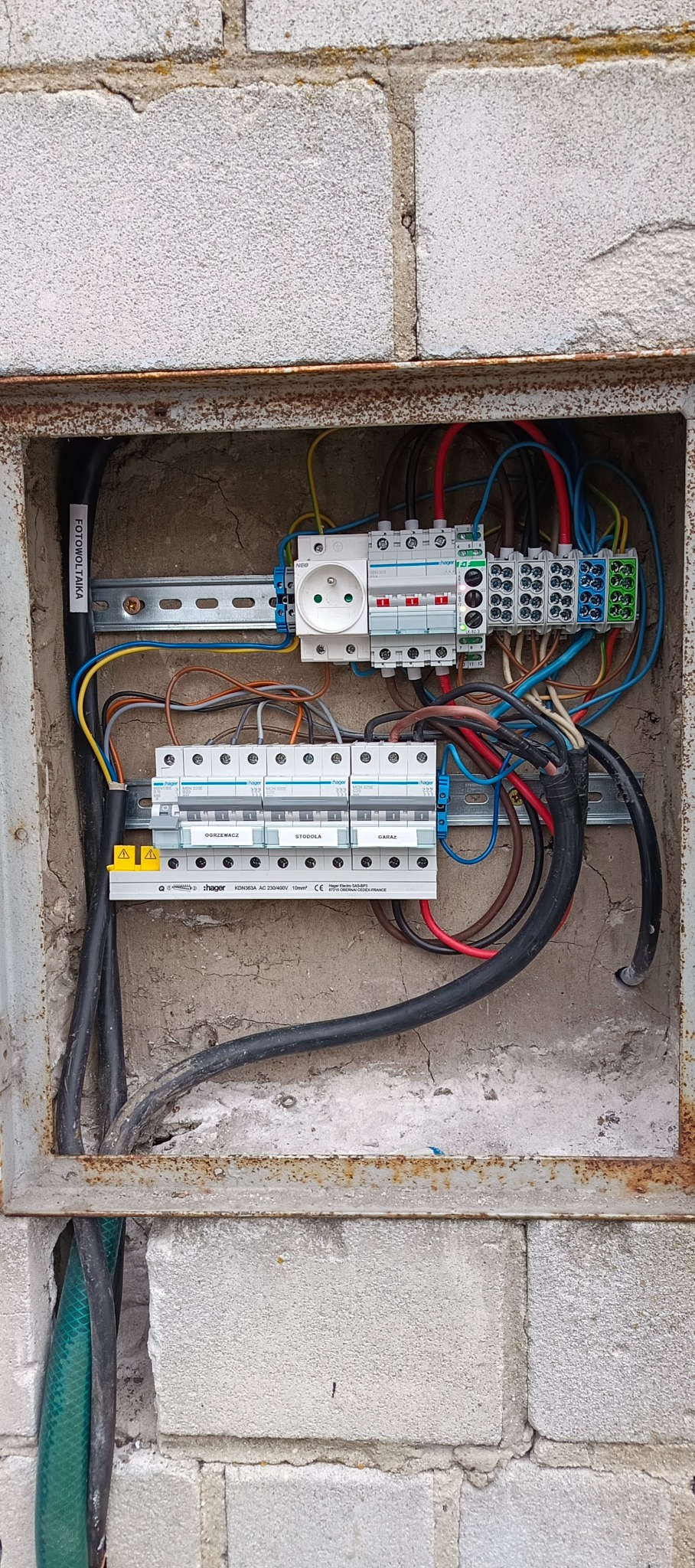 Rozdzielnica elektryczna Hager w obudowie z bloków betonowych. Widoczne bezpieczniki, gniazdo, przewody. Etykieta 'FOTOWOLTAIKA' na kablu. Oznaczenia: Ogrzewacz, Stodoła, Garaz.