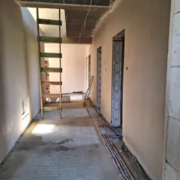 Wnętrze w trakcie prac wykończeniowych: świeżo otynkowane ściany w beżowym odcieniu, drewniana drabina, instalacja wzdłuż ściany i surowy betonowy strop.