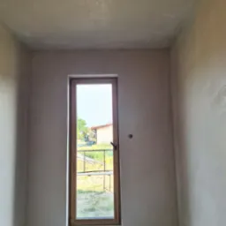 Wnętrze pokoju po wstępnym wyrównaniu ścian, widoczne szare tynki i drewniane okno balkonowe z widokiem na zewnątrz. Na podłodze widoczne instalacje.