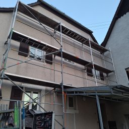 PerfectTynk - Elewacja domu w trakcie renowacji z użyciem rusztowania. Widoczna skrzynka z narzędziami i fragment drugiego budynku obok. Czyste niebo w tle.