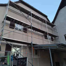 Elewacja domu w trakcie renowacji z użyciem rusztowania. Widoczna skrzynka z narzędziami i fragment drugiego budynku obok. Czyste niebo w tle.